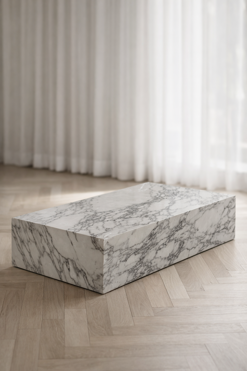 A minimal rectangular block coffee table, with white marble and delicate grey veining in a living room with sheer curtain and herringbone flooring. Angled view. 