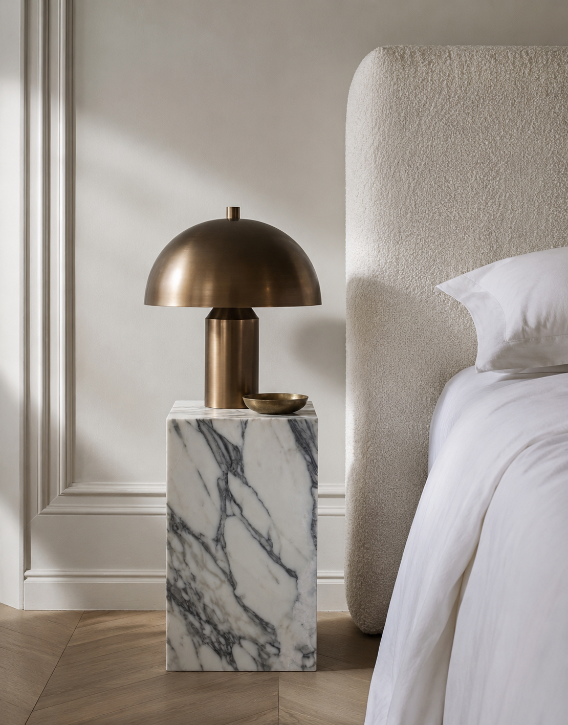 A bronze table lamp on a premium quality marble side table with white marble with grey veining and honed finishing, styled next to a white boucle bed with white linen beddings in a sun-lit Australian modern bedroom, highlighting the organic stone patterns.