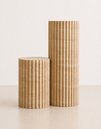 A pair of short and tall fluted beige travertine plinths with natural grain texture placed in a off white studio.