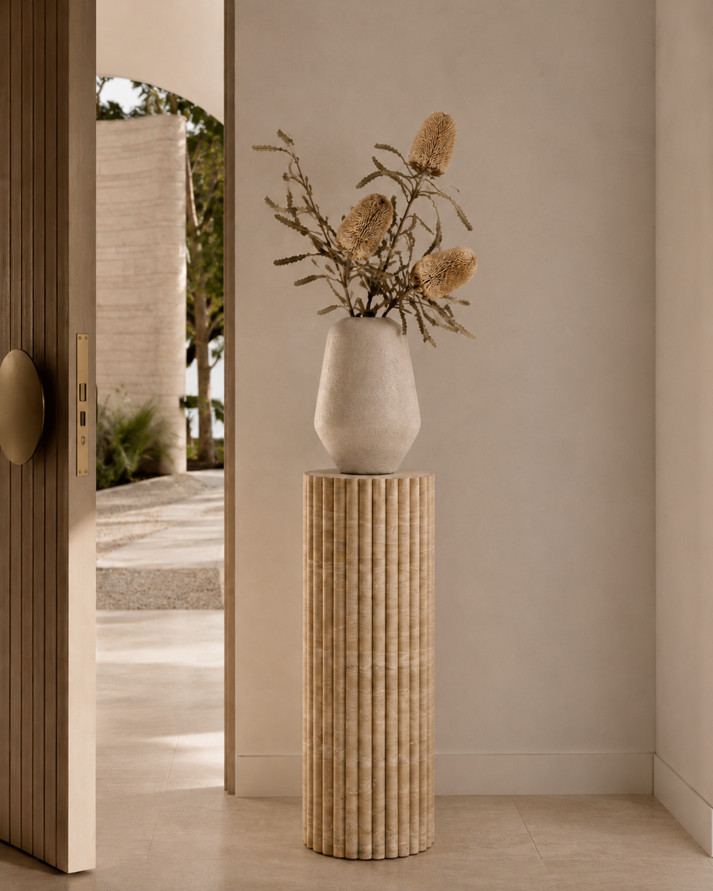Decorative vase with dried flowers on a beige travertine fluted pedestal in an entryway of a modern luxury Australia home next to a light timber door with brushed gold hardware.