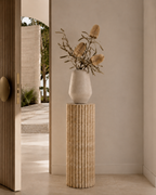 Decorative vase with dried flowers on a beige travertine fluted pedestal in an entryway of a modern luxury Australia home next to a light timber door with brushed gold hardware.