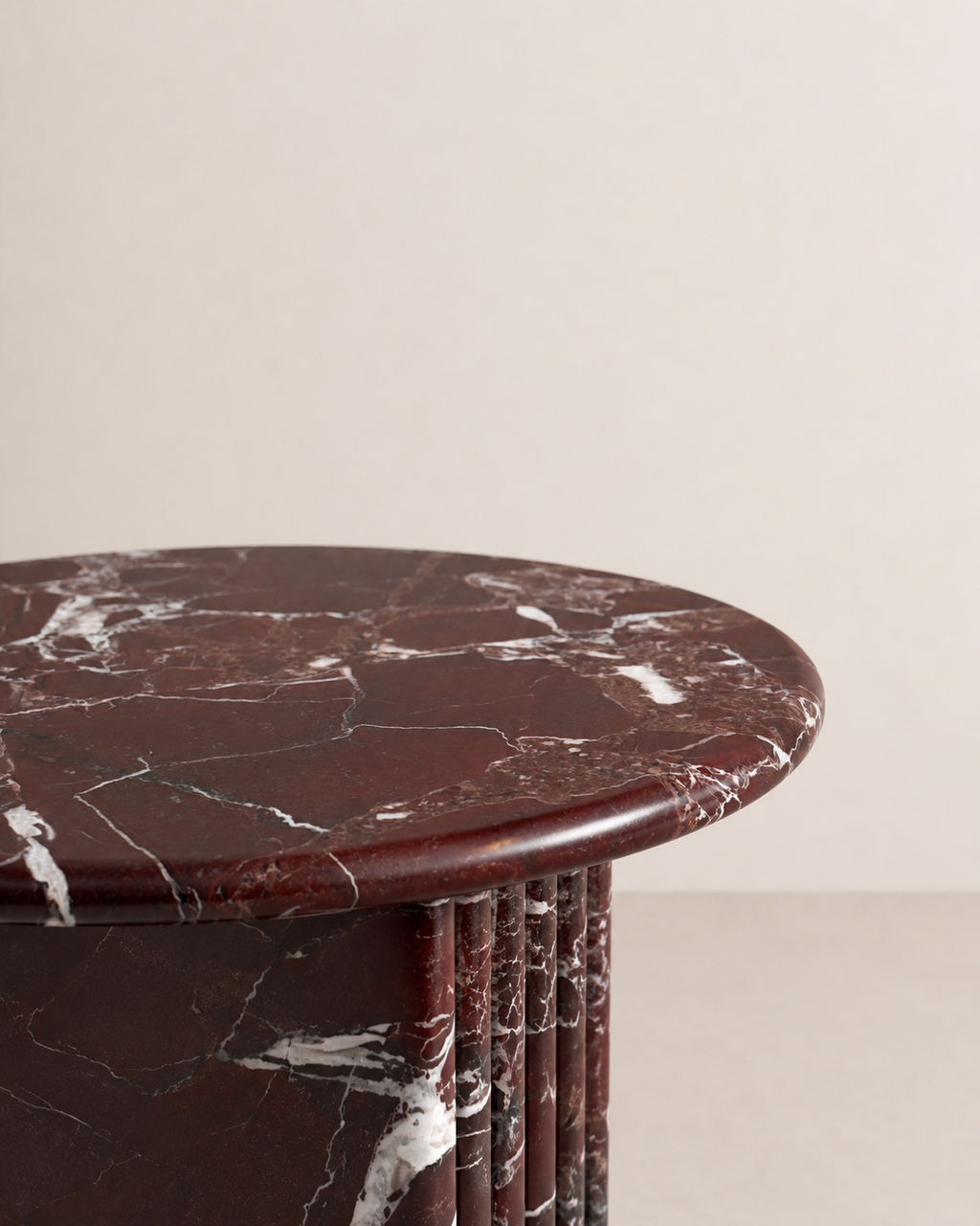 Close up of a luxury burgundy red marble side table with a ribbed cylindrical design in a beige background. Honed finishing.