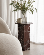 A luxury burgundy red marble side table with a ribbed cylindrical design  with a vase of greenery and a cup next to a white armchair. Modern Australian lifestyle