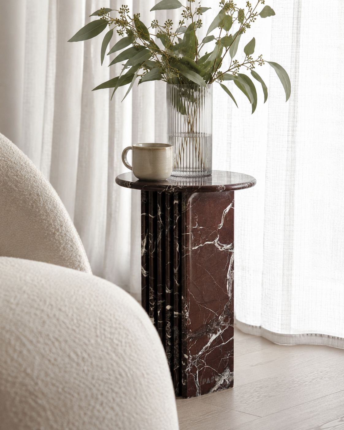 A luxury burgundy red marble side table with a ribbed cylindrical design  with a vase of greenery and a cup next to a white armchair. Modern Australian lifestyle
