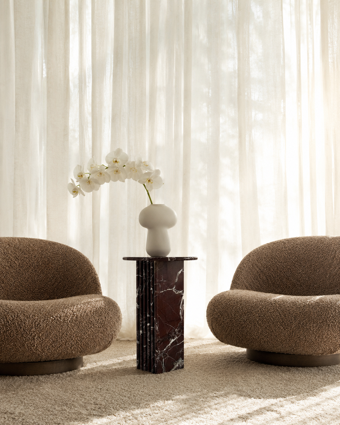 Two brown textured chairs with a luxury burgundy red marble side table with a ribbed cylindrical design  and white orchids in a room with sheer curtains.