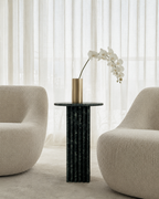 Deep emerald green marble side table with a ribbed cylindrical design, placed with a beige armchair and candle in a lounge room with curtains. Modern interior.