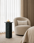 Deep emerald green marble side table with a ribbed cylindrical design, placed with a beige armchair and candle in a lounge room with curtains