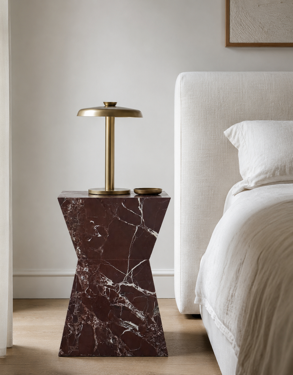 Hourglass shaped marble side table, made of luxury burgundy red marble with delicate white veining. A gold lamp placed atop of the table, sits next to a white linen bed in a aesthetic Australian bedroom.