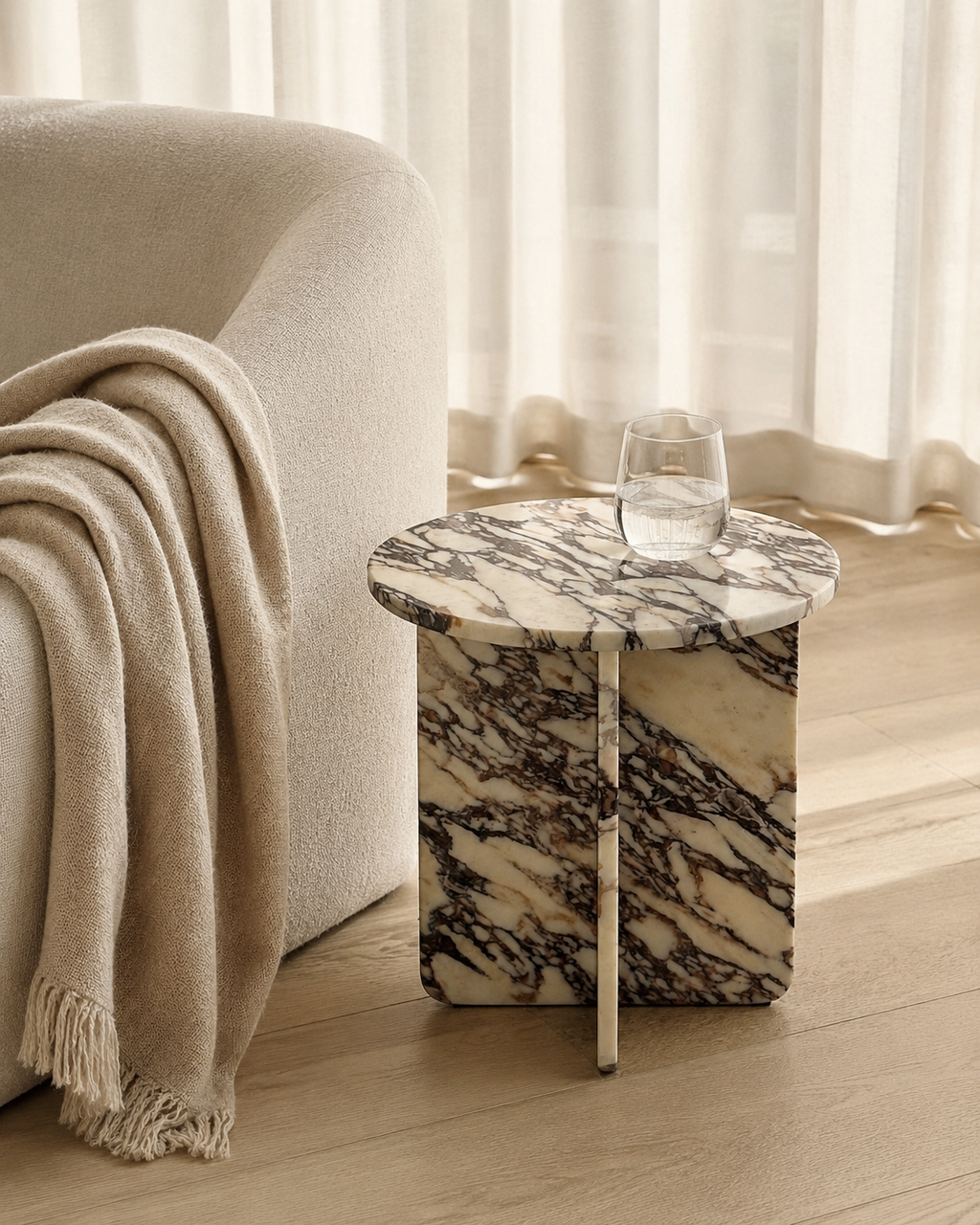 A luxury cream marble with delicate violet veining pattern round side table  with a glass of water next to a beige armchair in a softly lit living room. Modern Australian lifestyle.