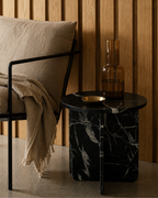 A luxury black marble with delicate white veining pattern round side table  next to a chair with a beige linen pillow and blanket.